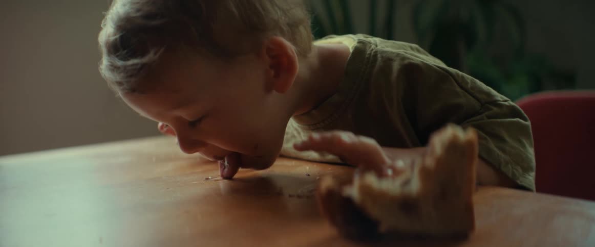 A baby licking peanut butter from a table while eating a sandwich at home