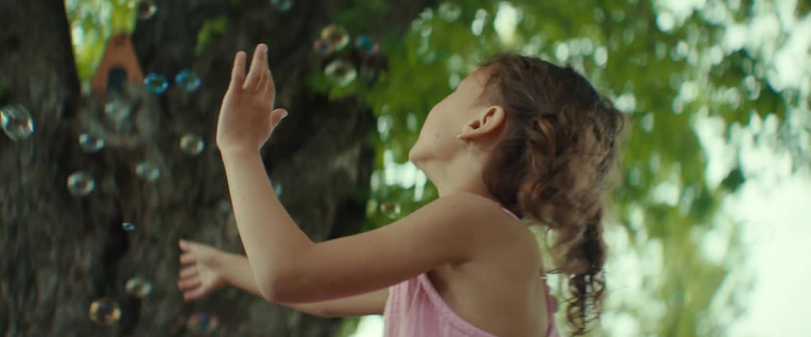 A little girl is outside under a tree chasing and popping bubbles