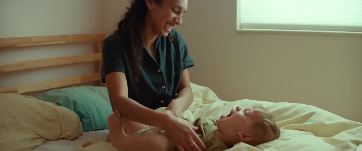 A mother laughing while tickling her smiling baby on a bed