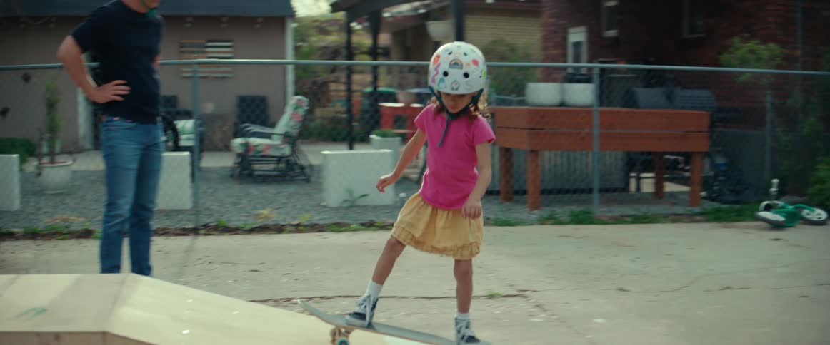 A little girl wearing a helmet rides a skateboard up a ramp and turns around going back down the ramp as her father watches