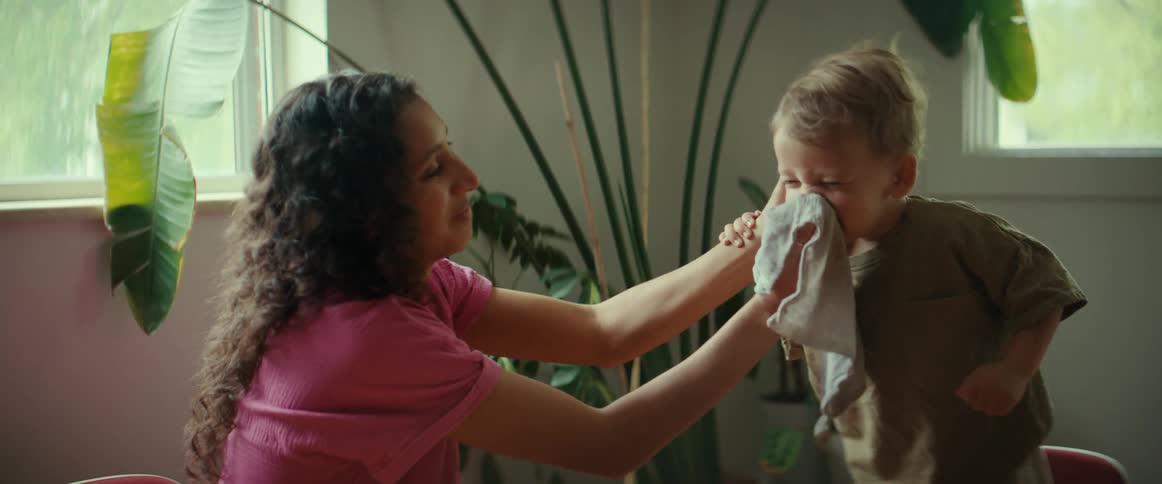 Mother wipes face of son as she sits near windows and large potted plant