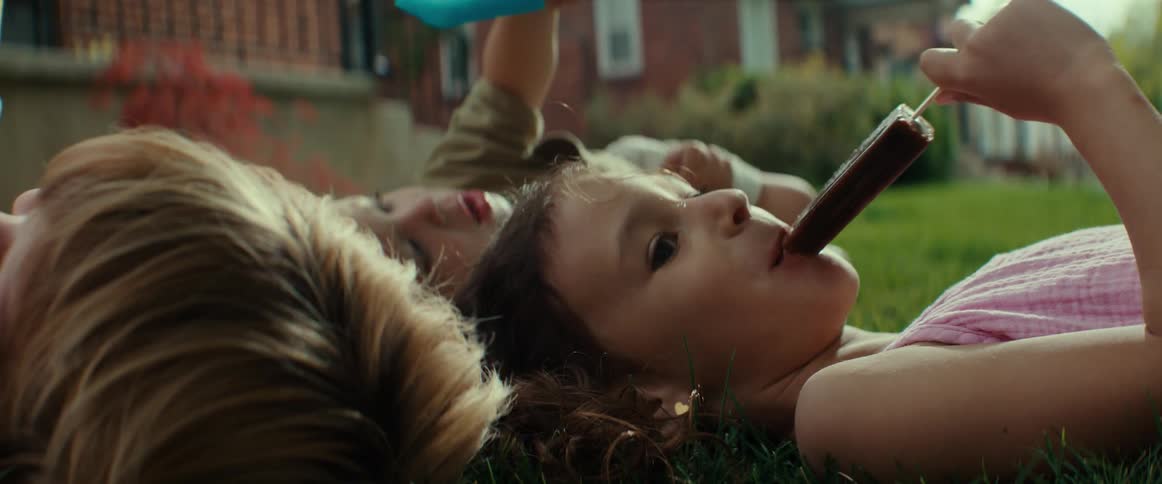 A little girl and two little boys lying on the front yard grass head to head eating popsicles