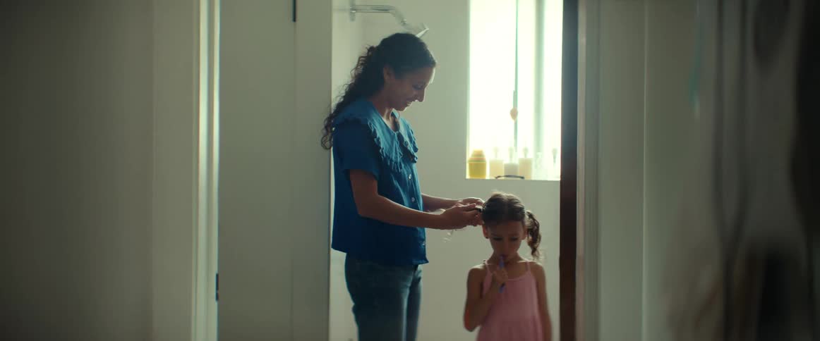 A mother and daughter stand in the doorway of a bathroom as the mom does the childs hair and the girl brushes her teeth