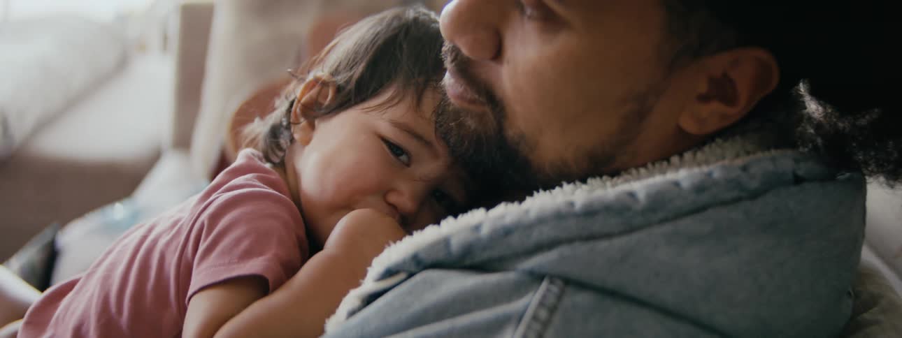 A young girl rests against the chest of her father indoors during the day