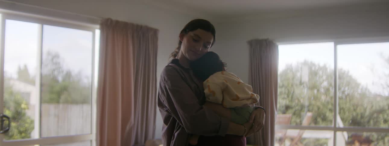 A pregnant woman holds a sleeping child in her arms while standing in a house during the day