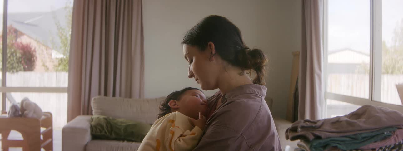 A woman rocks a sleeping child in her arms while sitting in the living room of a house during the day