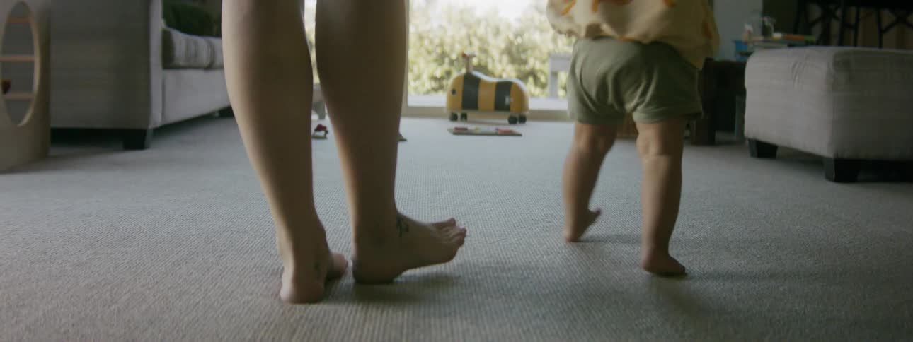 A woman helps her young child as he walks across a room at home