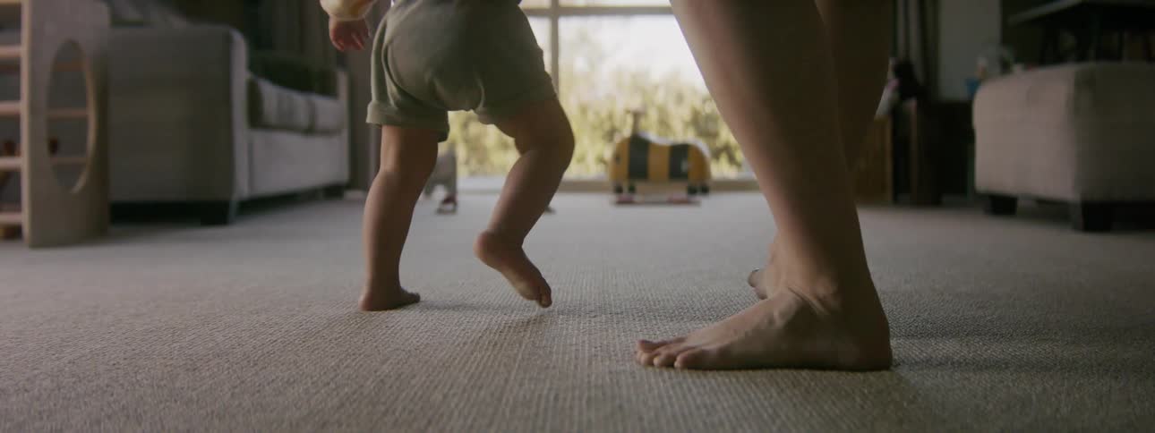 A mother sets her baby down and follows them as they walk across a room