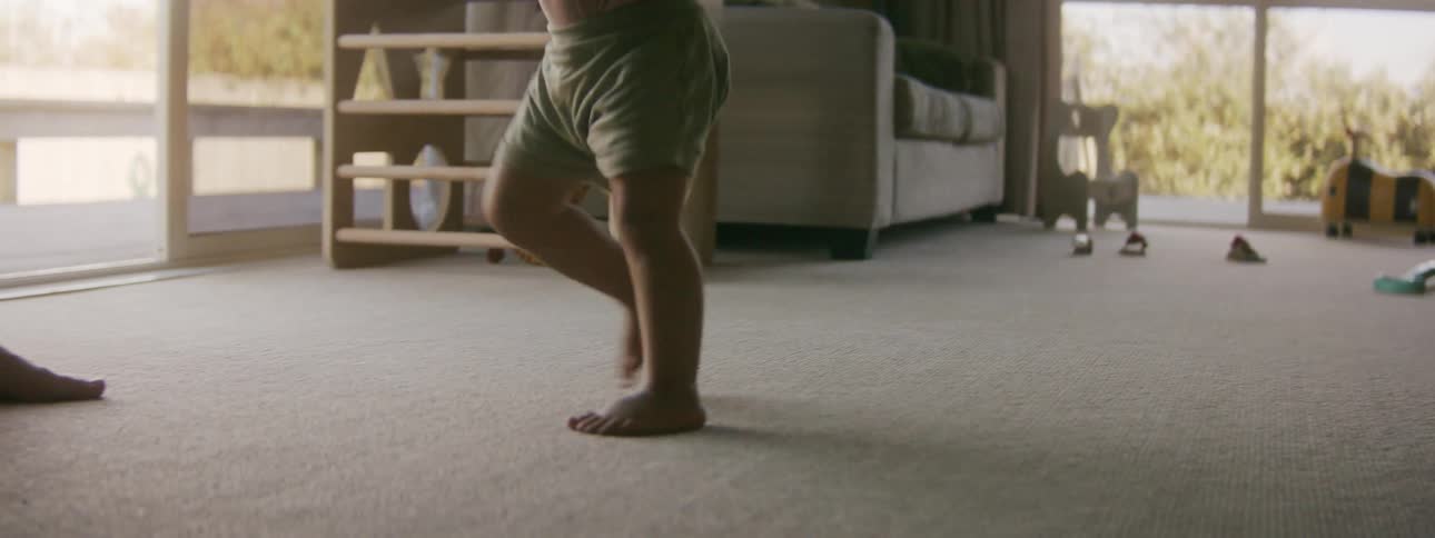 A small child walks across the living room of a house to approach an adult standing on the other side