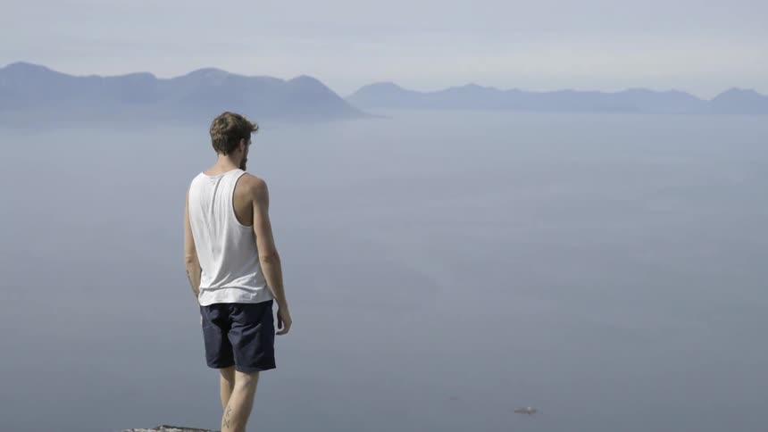 A man in a white shirt walks to the edge of a steep mountain cliff.
