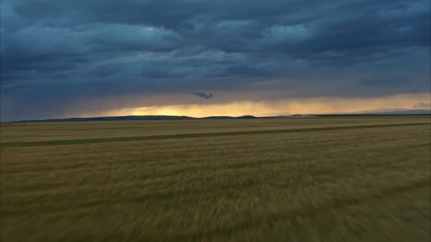 Crops stretch across land to mountains and bright horizon under dark storm clouds
