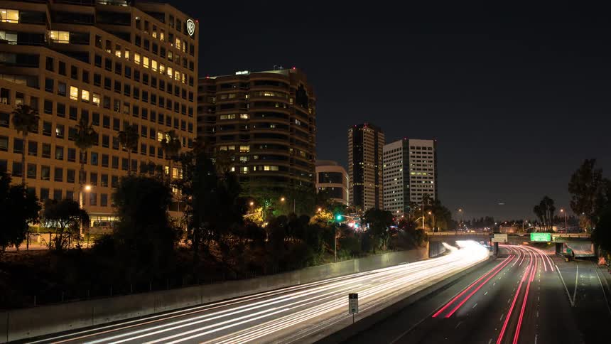 Headlights and taillights create a continuous stream of color on a highway near city buildings