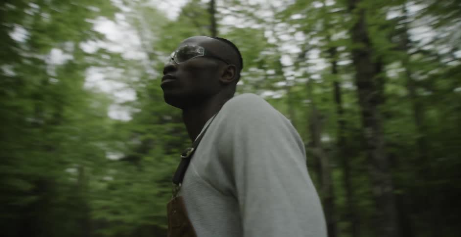 Black man wearing eye glasses and overalls stands in the forest
