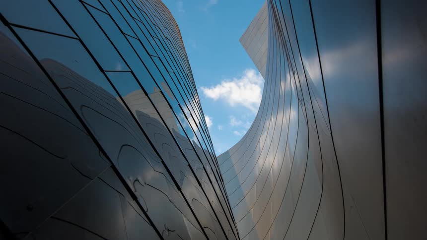 Clouds moving across a blue sky are seen above the reflective curved walls of a building