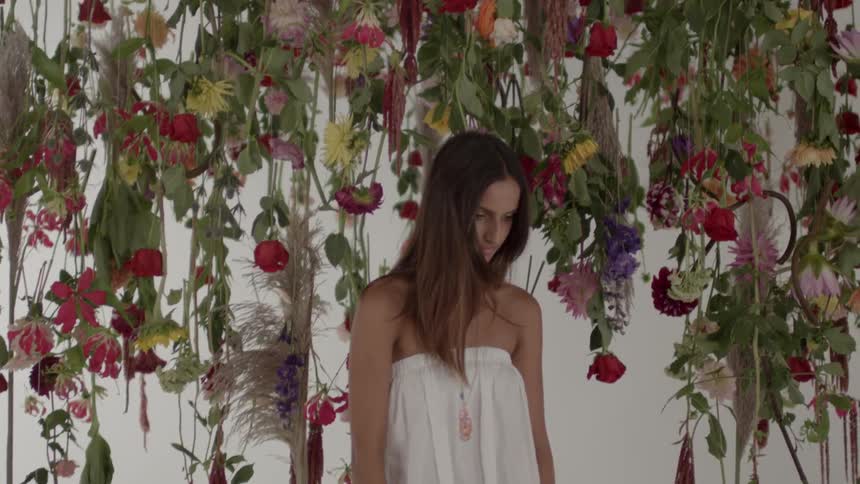 A young woman in a white dress stands amidst hanging flowers and briefly looks at the camera