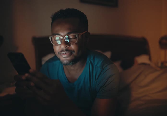 Man looks at a cellphone and smiles while sitting on the edge of a bed as a woman sleeps in the bed