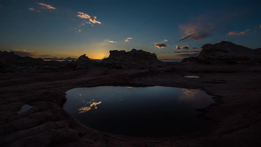 Sun sets and night falls as moving clouds are reflected in pool in stone ground surrounded by outcroppings that become silhouettes against red sky
