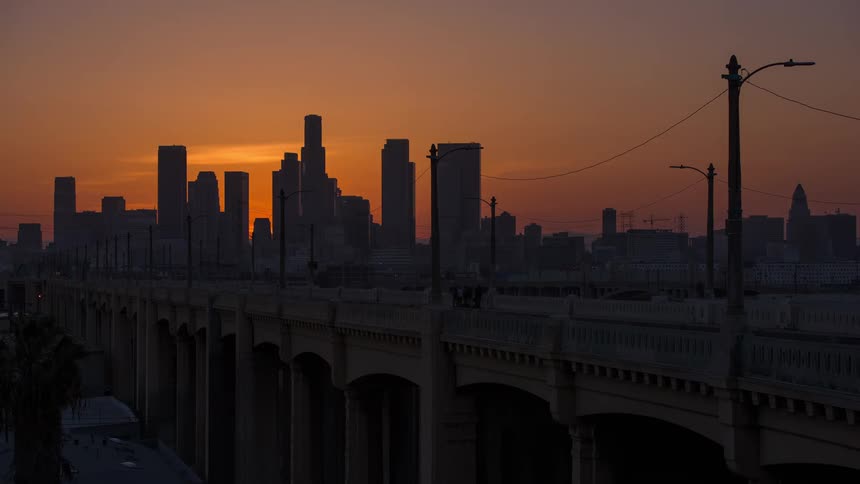 The sun sets behind the silhouette of city buildings while cars and people travel across a bridge
