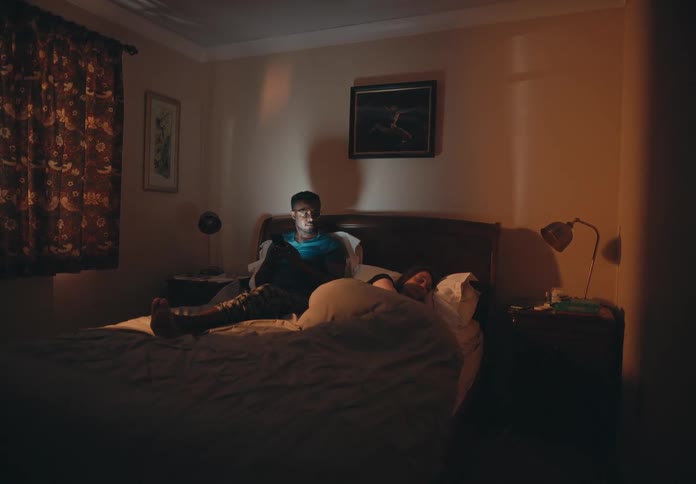 A man sitting up in bed using his phone turns his head and looks at his sleeping wife