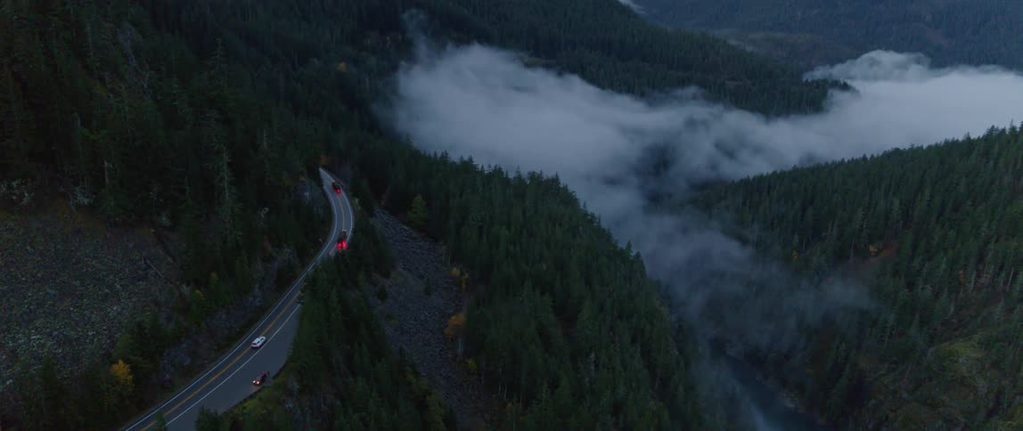 Vehicles drive down a narrow mountain road in heavy fog