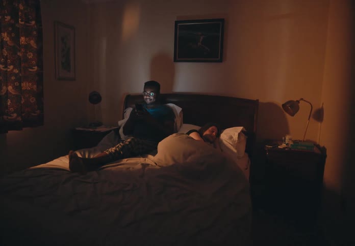 A woman sleeps in bed while her husband sits up on his smartphone