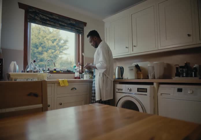 A man standing in the kitchen pouring coffee into a mug while holding his phone and texting