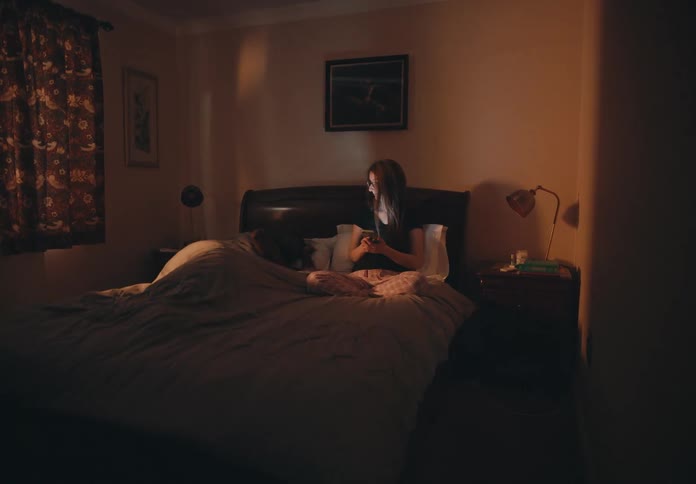 A woman sitting up in bed texting on her phone looks over at her sleeping husband