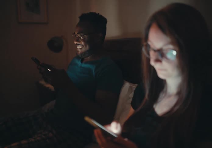 A couple sits side by side in bed ignoring each other as they look at their smartphones