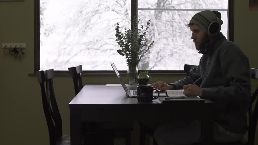 A man sitting at a table in a cafe working on a laptop as snow is seen falling outside