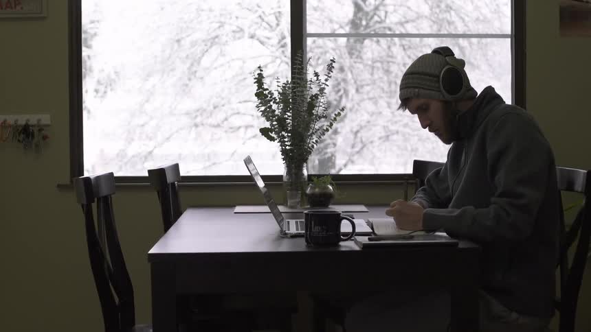 A man wearing headphones studies at a table as snow falls outside the window
