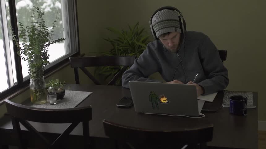A man sits at a table writing and working on a laptop