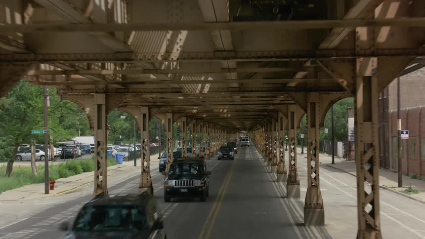 Cars traveling on a city street under the elevated train tracks near a public park