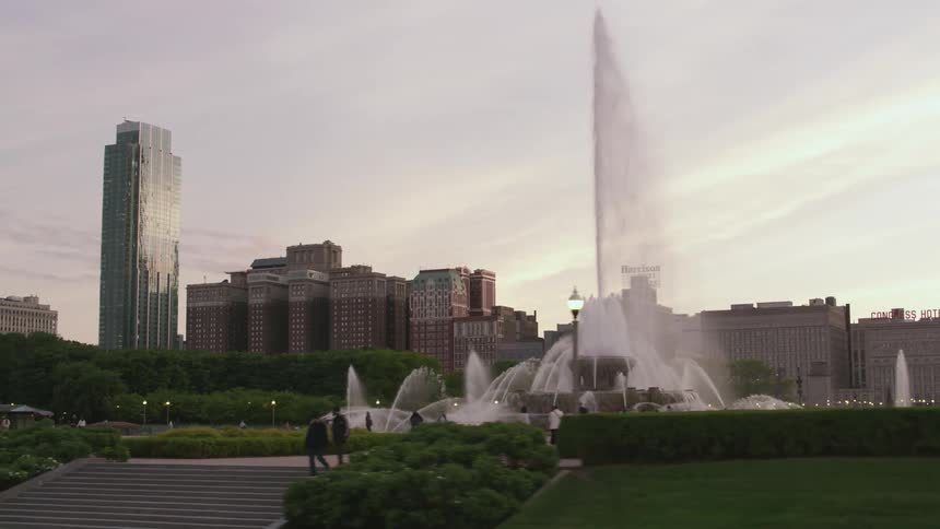 Tall buildings are on the city skyline traveling by a park with people walking near a fountain