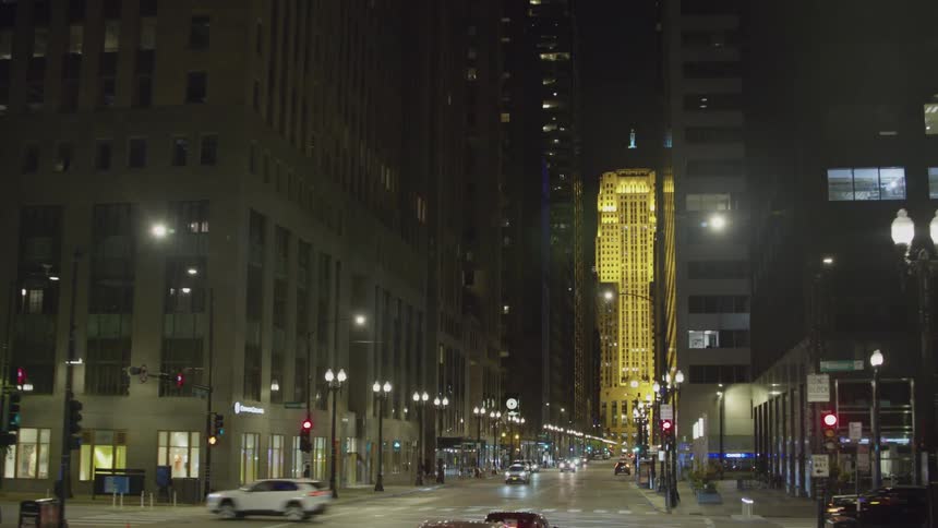 A city street with high rise office buildings with cars driving and a pedestrian walking at night