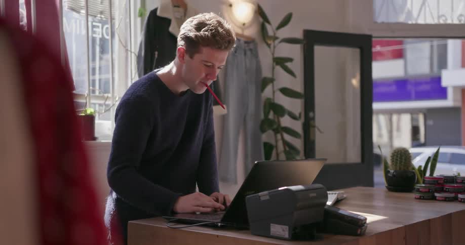 A man in a clothing store types on a laptop behind the counter