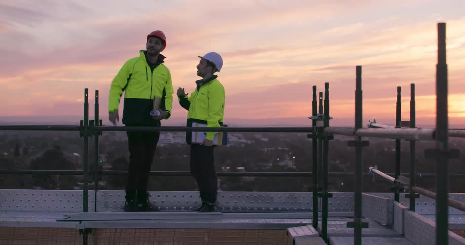 Two men in hard hats stand and talk on a rooftop construction site