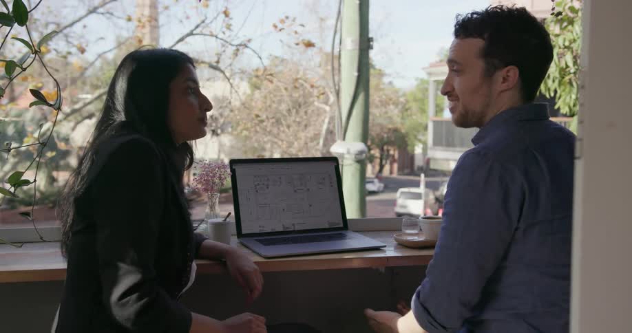 Two people are sitting in front of a window having a conversation with a laptop in front of them