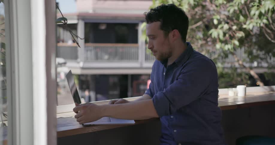 A man leans by the counter using his phone and laptop