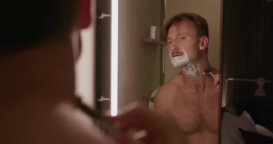 A man looks in a bathroom mirror and shaves his beard