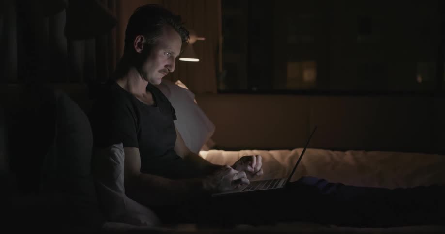 A man sits in his bed using a laptop at night
