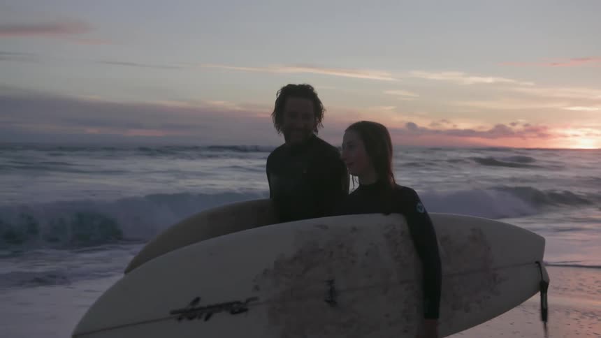 Man and woman carrying surfboards walk on the beach at sunset