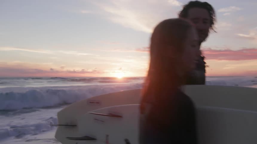 Two people walk from the water with surfboards