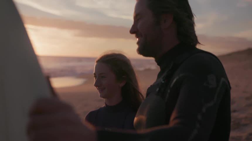A man and a young girl are each holding surfboards on the beach at sunset