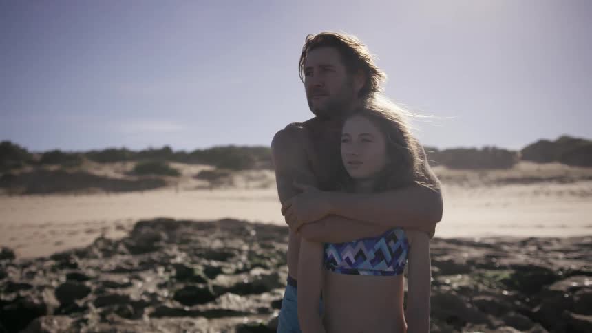 A man is hugging his daughter at the beach and points to something off in the distance
