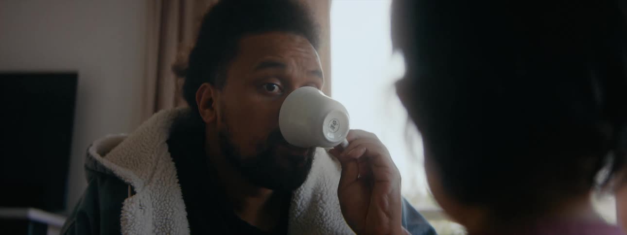 A man drinking out of a coffee cup listens to another man mostly offscreen