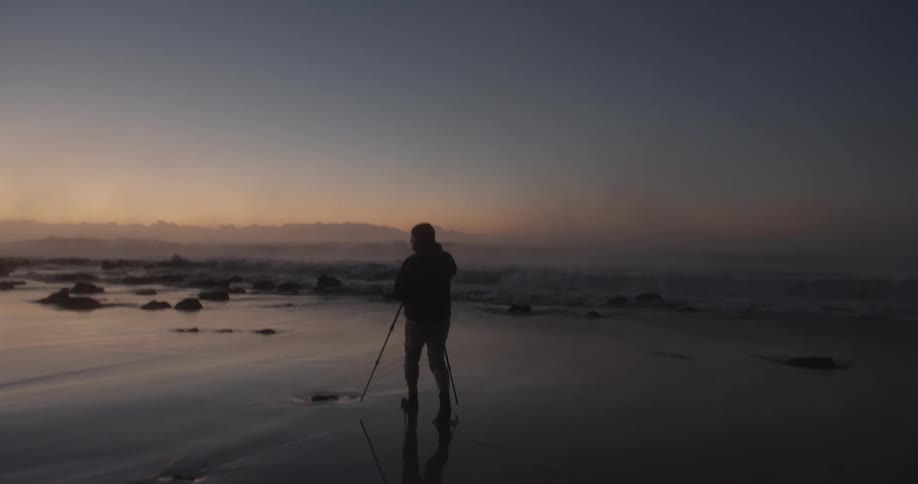 A man is walking on a wet beach with a tripod and camera as the sun is low on the horizon