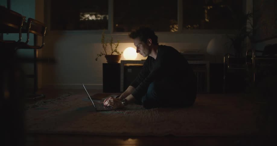 A man is sitting on a rug in a living room and typing on a laptop