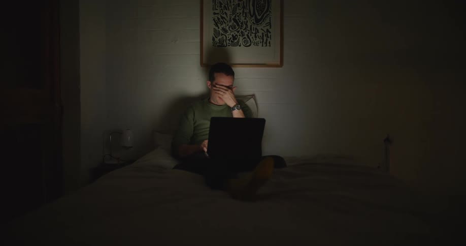 Man with mustache and green sweatshirt sitting on bed yawns and closes laptop in room at night