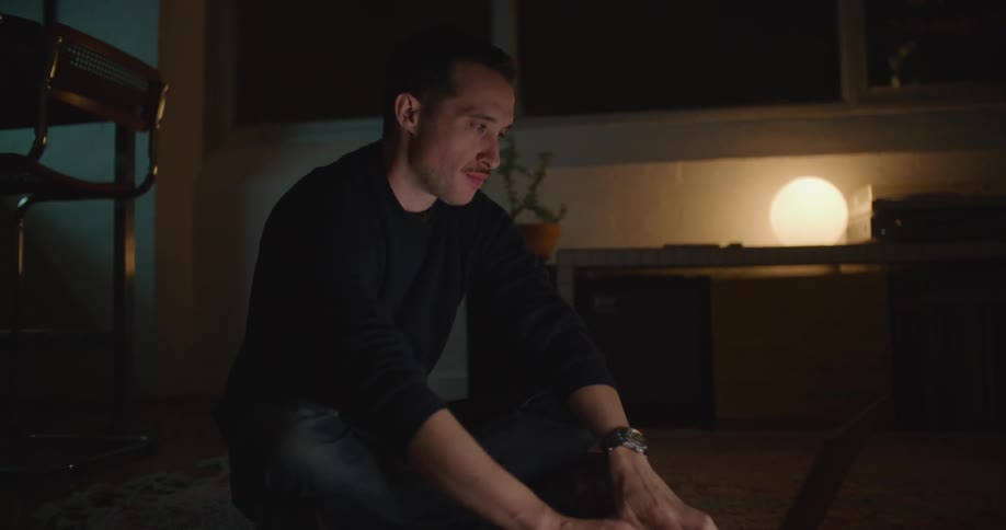 Man with mustache green sweatshirt and jeans sits on floor working on laptop next to dining table at night