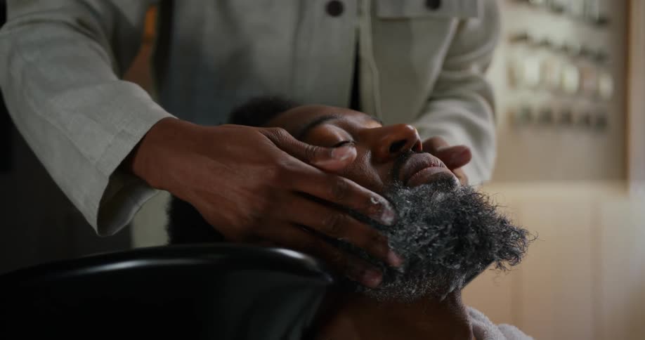 A man is sitting in a chair and another man wearing a white coat is massaging his face and beard